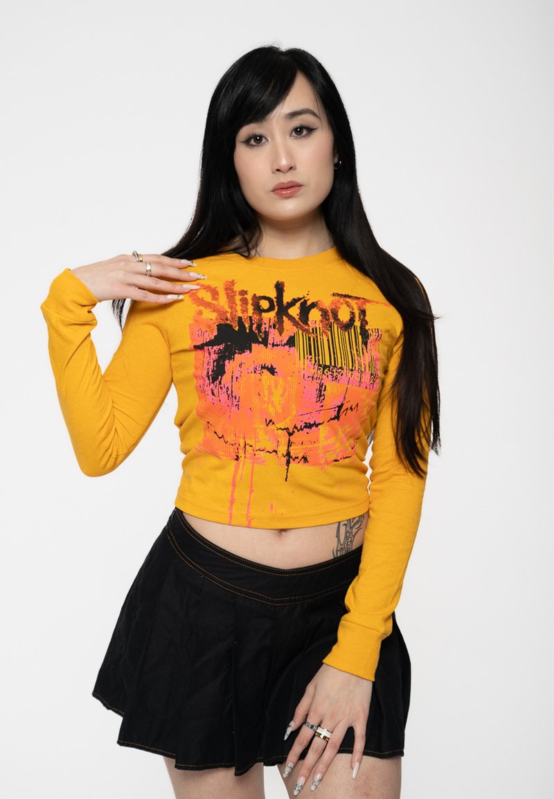 Yellow long-sleeve crop top featuring black and pink graphic print with drips, paired with a black pleated mini skirt.