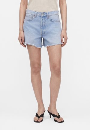 Kobieta w jasnoniebieskich szortach jeansowych z nieobrobionym dołem i czarnych sandałach na obcasie, stojąca na białym tle.