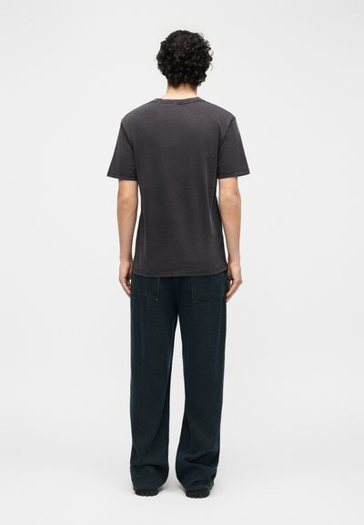 Άτομο με σγουρά μαλλιά στέκεται με την πλάτη, φοράει σκούρο γκρι κοντομάνικο πουκάμισο και άνετο μαύρο παντελόνι σε απλό φόντο.