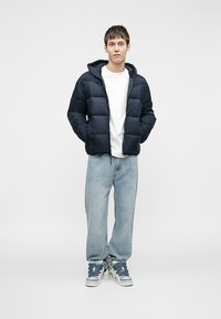 Joven con una chaqueta de plumas negra, camiseta blanca, jeans de mezclilla azul claro y zapatillas azul y blancas, de pie con las manos en los bolsillos.