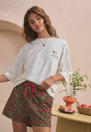 Bela oversize majica s grafiko napitka in kratkimi hlačami z leopardjim vzorcem, okrašenimi z rdečimi češnjami, dopolnjenimi z rdečo vrvico.