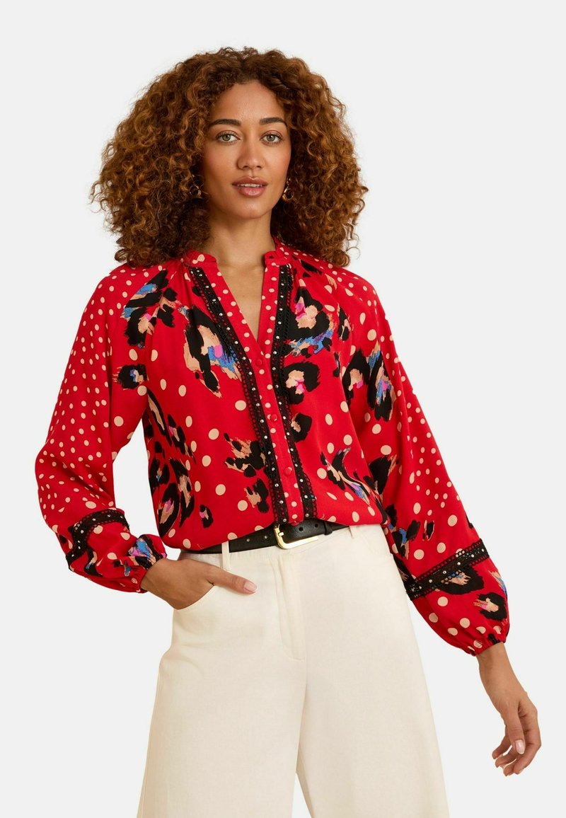 Sarkana blūze ar punktiem un ziedu rakstu. Ar pogu aizdari priekšpusē un melniem akcentiem, ar pūkainām garām piedurknēm. Valkāta kopā ar baltām platām biksēm.