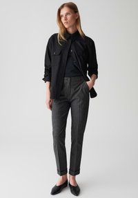 Zwarte overhemd met knopen over een zwarte top, gecombineerd met grijze, gepatterneerde tailor pants. De broek heeft een omslagzoom en zakken. Zwarte flats.