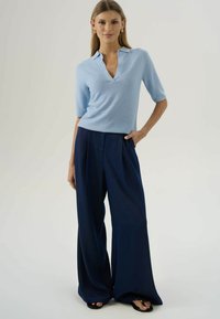 Žena s rovnými vlasmi v svetlomodrej blúzke s krátkym rukávom a širokými nohavicami námorníckej farby stojí s jednou rukou v vrecku na neutrálnom pozadí.