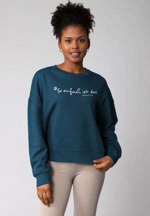 Teal oversized sweatshirt met een ronde hals en geribde mouwen, voorzien van witte tekst "So einfach ist das" over de borst.