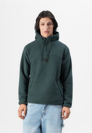 Pull en polaire vert avec col haut, cordon de serrage réglable et fermeture éclair. Comprend deux poches latérales et un patch logo sur la poitrine.