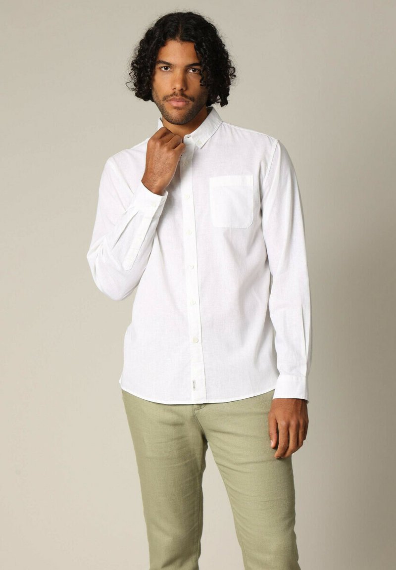 Witte blouse met lange mouwen en een enkele borstzak, met een licht gebogen onderkant, gecombineerd met lichtgroene broek.