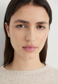 Portret van een persoon met middengroot bruin haar, een gladde huid, zachte roze lippen en neutrale make-up, gekleed in een licht textuurtrui.