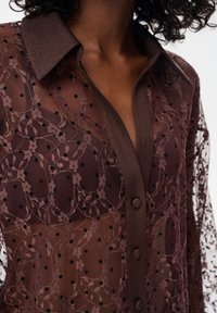 Przezroczysta brązowa koszula z kołnierzykiem zapinanym na guziki, ozdobiona kwiatowymi wzorami z koronki oraz czarnymi kropeczkami. Teksturowany materiał z delikatnym połyskiem.