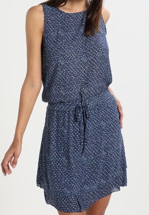 Kobieta ubrana w granatową sukienkę bez rękawów z wzorem i sznurkiem w talii, pozująca z jedną ręką na biodrze.