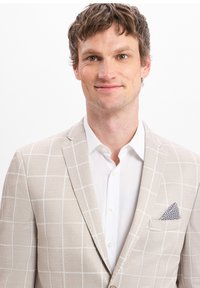 Mann mit kurzen lockigen braunen Haaren, der ein beiges karierter Blazer über einem weißen Hemd mit einem gemusterten Taschentuch trägt.