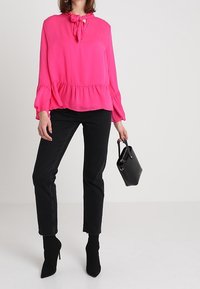 Blusa fucsia con scollatura a fiocco, orlo plissettato e maniche lunghe; abbinata a jeans neri a vita alta e stivaletti con punta.