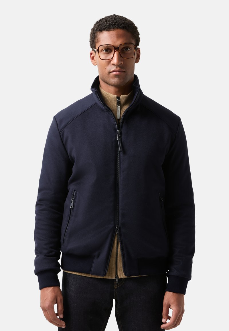Mann in einer marineblauen Zip-Jacke über einem beigen Zip-Pullover und dunklen Jeans, der vor einem schlichten hellen Hintergrund steht.