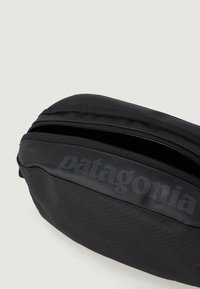 Černá zipová taštička vyrobená z texturovaného syntetického materiálu. Obsahuje výrazné tištěné logo. Kompaktní oválný tvar s jemnými švy.