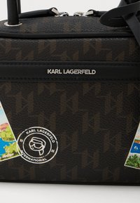 Ciemnobrązowa torba Karl Lagerfeld z wytłoczonym wzorem, srebrnym logo, naklejkami w tematyce podróżniczej oraz widoczną na górze rączką zamka błyskawicznego.