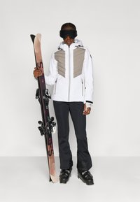 Biely lyžiarsky kabát s béžovou prešívanou hornou časťou, čierne nohavice a lyžiarske topánky. Lyže držané v ľavej ruke, na hlave lyžiarske okuliare.