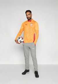 Narančasta sportska majica dugih rukava s teksturiranim uzorkom i crvenim detaljima; sive trenirke; osoba drži crno-bijelu nogometnu loptu; crne tenisice.