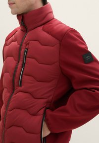 Czerwona ocieplana kurtka z wysokim kołnierzem, pikowaniem w zygzak na przodzie, siateczkowymi wstawkami na ramionach oraz zamkiem błyskawicznym w kieszeni na piersi.