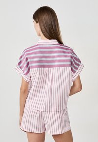 Femme avec de longs cheveux bruns portant une chemise à col à manches courtes, boutonnée, à rayures horizontales et verticales mélangées, ainsi que des shorts à rayures assorties.