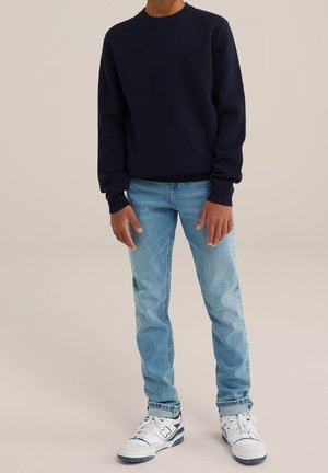 Dziecko w granatowym swetrze, jasnoniebieskich dżinsach i białych sneakersach stoi na jednolitym tle.