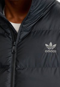 Svart uppblåst väst med hög krage, dragkedjestängning, och en broderad Adidas-logotyp på vänster bröst. Slät, glänsande tygstruktur.