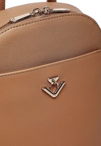 Hnědý kožený batoh s zaobleným vrchem, který je vybaven stříbrným zipem a výrazným kovovým logem na přední kapse.