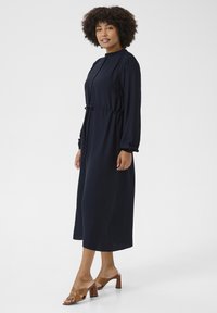 Námornícky modrá dlhá šaty s dlhým rukávom a predným zapínaním na gombíky, so sťahovacím pásom a nariasenými rukávmi; v kombinácii s hnedými sandálmi na blokovom podpätku.