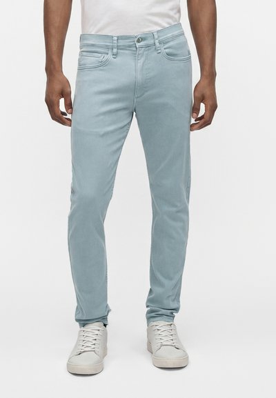 Homme portant un jean slim bleu clair et des baskets blanches, debout devant un fond clair uni.