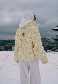 Osoba v bielej pletenej mikine s kapucňou a krémovom hrubom pletenom svetri stojí na zasneženom povrchu s oblačnou oblohou a vzdialenými kopcami.