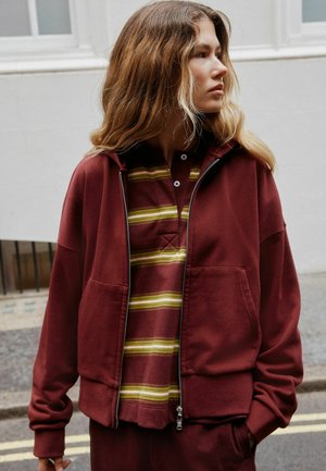 Junge Person mit langen, welligen Haaren, die eine bordeauxrote Jacke mit Reißverschluss über einem gestreiften bordeauxroten und gelben Hemd trägt und im Freien seitlich schaut.