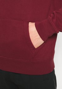 Sudadera burdeos con bolsillo tipo canguro, puños y dobladillo acanalados. Textura suave con un diseño ajustado y elementos elásticos.
