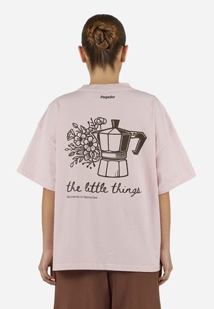Person mit einem Dutt, der ein hellrosa Hemd trägt, auf dem Rücken eine Kaffeekanne, Blumen und der Text "the little things Moments to Memories".