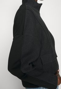 Maglione nero con collo alto, caratterizzato da una trama a spina di pesce nera su un lato e tessuto nero uniforme sull'altro. Maniche lunghe e tasca frontale.