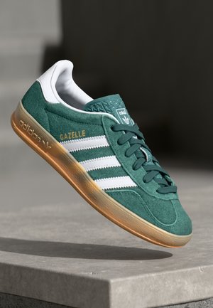 Baskets Adidas Gazelle vertes avec des bandes blanches et une semelle gomme flottant au-dessus d'une surface en béton gris.