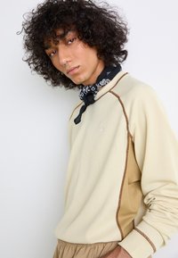 Beige ribbed sweatshirt with brown accents and textured side panels. Features a high neckline and fitted cuffs. Black bandana around the neck.