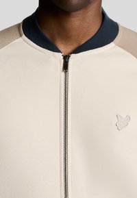 Beige zip-up jakke med en marineblå krave, med lysebrune skulderpaneler og et lille broderet logo på brystet. Blød tekstur.