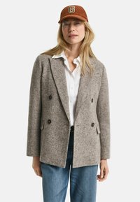 Blazer grigio in misto lana doppiopetto con due ampi taschini anteriori e bottoni scuri, abbinato a una camicia bianca e jeans in denim.