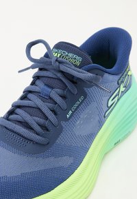 Skechers løpesko med en blå mesh-overdel, grønn såle med detaljer og lisser. Merking synlig på siden og tungen. Holdbar og polstret design.