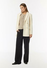 Double-breasted cream blazer with vertical stripes, paired with a beige ribbed sweater and loose black trousers. Black flat shoes complete the look.