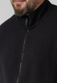 Zwarte zip-up jas van zacht materiaal, met een hoge kraag en een gladde ritsafwerking, en een licht gestructureerde uitstraling.