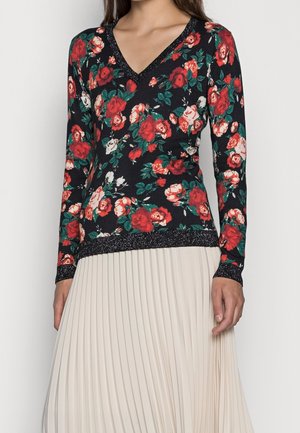 Femme portant un haut noir à manches longues avec imprimé floral rouge et blanc et bordure scintillante, associé à une jupe plissée beige clair.
