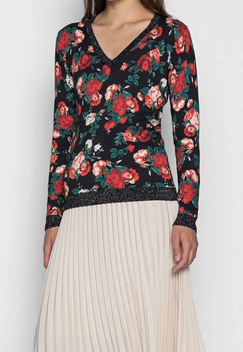 Femme portant un haut noir à manches longues avec imprimé floral rouge et blanc et bordure scintillante, associé à une jupe plissée beige clair.