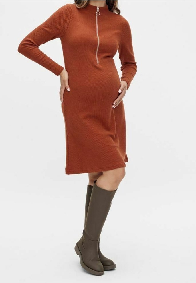 Femme en robe longue manche couleur rouille avec fermeture éclair frontale, tenant son ventre de femme enceinte, portant des bottes hautes marron foncé.