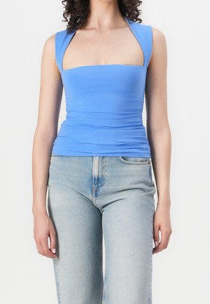 Nainen, jolla on päällään hihaton sininen tiukka toppi leveillä olkaimilla ja vaaleansiniset korkeavyötäröiset farkut, seisoo yksinkertaisen taustan edessä.