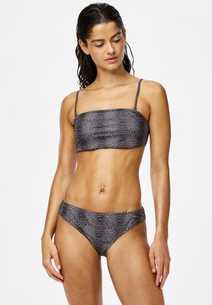 Žena nosi dvojdielne leopardie plavky s tenkými ramienkami, pozerá sa dole, na jednoduchom bielom pozadí.