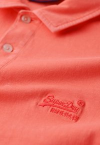 Koraalrode polo shirt van katoen, met een kraag, twee knopen en een geborduurd "Superdry"-logo op de borst. Soepele textuur.