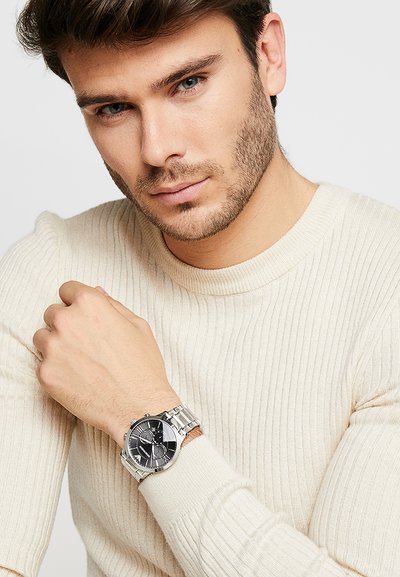 Reloj de hombre con una correa de metal plateado, esfera negra y acentos plateados. Llevado en una muñeca contra un suéter de punto beige claro.
