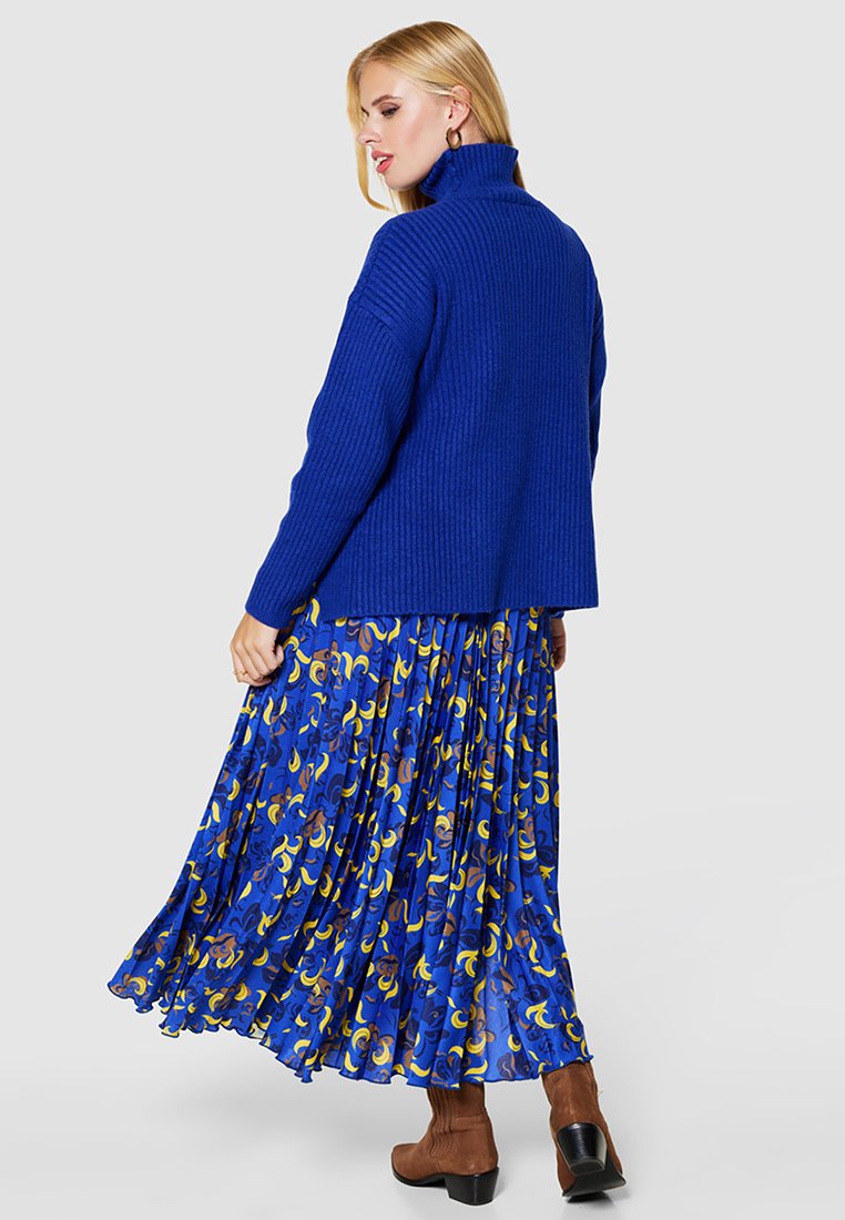 Szafranowo-niebieski sweter z golfem z prążkowanego materiału, połączony z lejącą się, plisowaną spódnicą o żywym, abstrakcyjnym wzorze w odcieniach niebieskiego, brązowego i żółtego.