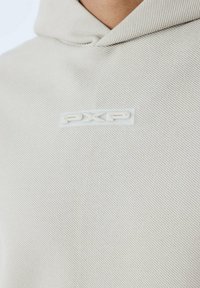 Felpa beige con tessuto testurizzato, con un piccolo patch logo trasparente etichettato "PXP" sul davanti. Il design include un cappuccio con lacci.