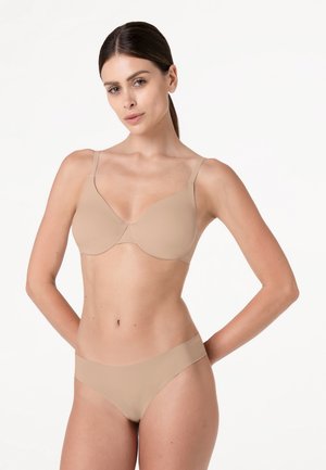 Donna con capelli scuri raccolti dietro, che indossa un reggiseno beige e biancheria intima abbinata, in piedi davanti a uno sfondo bianco uniforme.